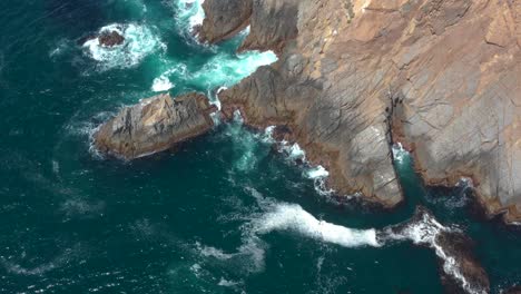 Waves-hitting-and-splashing-rocks-at-the-coast-of-a-blue-sea,-drone-shot