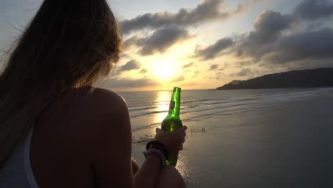 Chica-De-Pelo-Largo-Sentada-En-La-Playa-En-Una-Tarde-Ventosa-Con-Una-Cerveza-En-La-Mano-Y-Disfrutando-De-La-Puesta-De-Sol-En-Indonesia,-Isla-De-Lombok