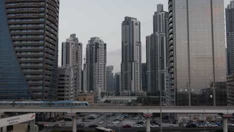 The-metro-train-passes-through-the-shot-in-slow-motion,-with-large-buildings-and-skyscrapers-in-the-background-in-the-middle-eastern-city-of-Dubai,-United-Arab-Emirates