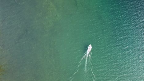 Bird's-eye-view-drone-shot-of-a-boat-slowly-cutting-through-the-beautiful-water-of-a-crystal-clear-lake-with-emerald-and-blue-hues-in-rural-northern-Canada