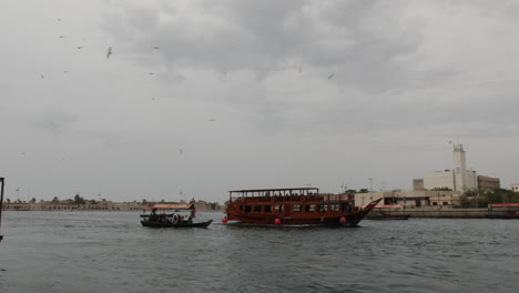 Nahöstliche-Boote,-Die-An-Einem-Bewölkten-Und-Bewölkten-Tag-Den-Fluss-In-Old-Dubai-Hinunterfahren,-Während-Möwen-In-Zeitlupe-Mit-60-Bildern-Pro-Sekunde-über-Ihnen-Fliegen