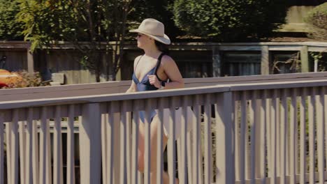 Glückliche-Frau-Mit-Bikini,-Sonnenbrille-Und-Sonnenhut,-Die-Auf-Einem-Holzsteg-Spazieren-Geht