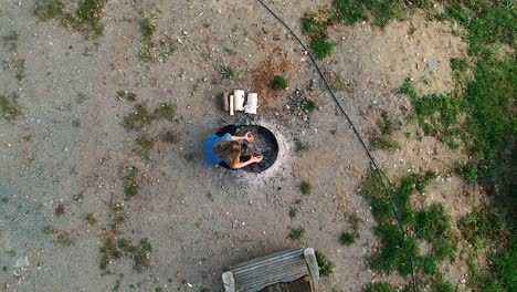 Unique-bird's-eye-view-perspective-of-a-young-women-walking-to-a-fire-pit-and-starting-to-stack-logs-into-a-teepee-to-prepare-for-a-fire-at-her-campsite-in-a-provincial-park-in-rural-Ontario,-Canada