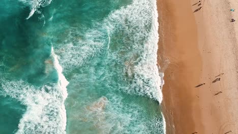 Top-down-birds-of-view-of-a-beautiful-tropical-beach-with-waves-crashing-into-the-sandy-shore-with-many-people-walking