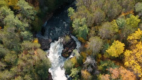 Upward-panning-drone-shot-of-a-waterfall-surrounded-by-peak-fall-foliage-in-Northern-Ontario,-Canada
