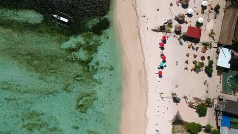 Bird's-eye-view-drone-shot-of-a-tropical-beach-with-colourful-umbrellas-and-a-canoe-just-off-the-shoreline-in-the-tropical-Gili-Islands,-Indonesia