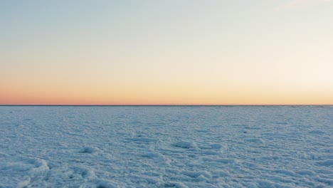 Low-and-quick-flying-over-big-white-field-of-crushed-pieces-of-ice-on-Baltic-sea,-Latvia-during-a-sunset