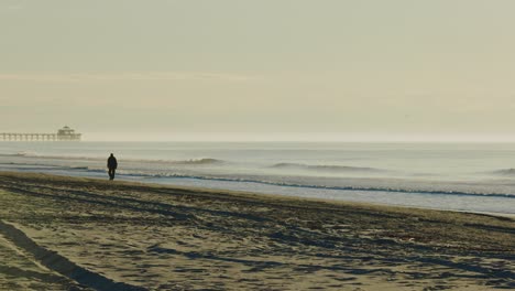 Cinematic-wide-shot-of-man-walking-along-sandy-beach-early-in-the-morning-during-sunrise
