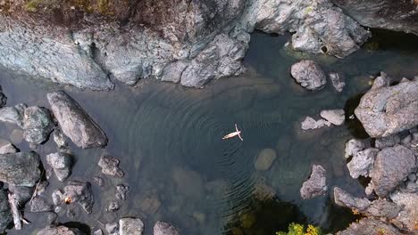 Top-down-bird's-eye-view-drone-shot-of-a-girl-in-a-bikini-floating-in-crystal-clear-water-in-a-rock-pool-located-in-the-country-side-of-British-Columbia,-Canada