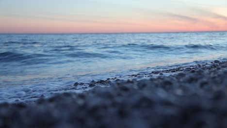 Sea-waves-splashing-on-shore-with-pebbles-at-twilight-reflecting-colorful-sky,-slow-motion