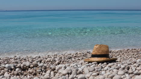 Straw-hat-left-over-empty-pebbles-beach-washed-by-emerald-crystal-sea-water,-holidays-concept