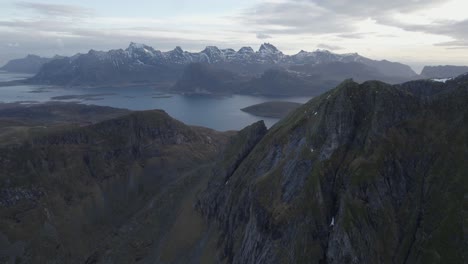 Aerial-view-of-steep-mountains-with-snowy-peaks-in-the-background,-in-Lofoten,-Norway---reverse,-tilt,-drone-shot