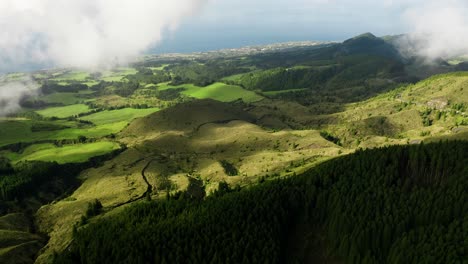 Aerial-push-out-over-pastures-and-forest-patches,-São-Miguel-shoreline-backdrop,-Azores