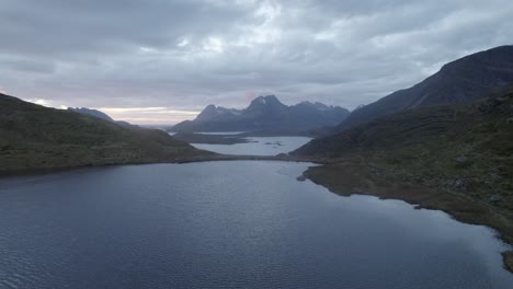 Aerial-view-low-over-a-pond-with-mountain-background,-gloomy-day-in-Lofoten,-Norway---pull-back,-drone-shot