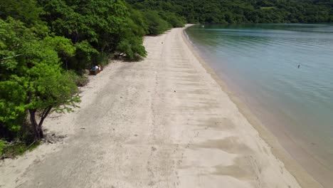 Aerial-dolly-in-of-couples-walking-in-sand-shore-between-dense-woods-and-sea-in-Nacascolo-beach,-Papagayo-Peninsula,-Costa-Rica