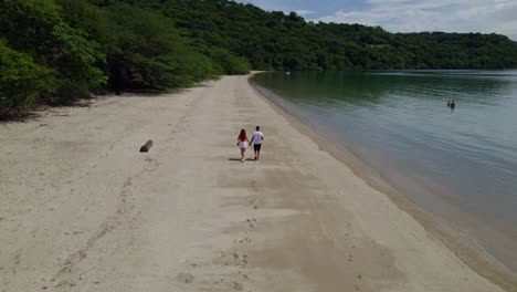 Aerial-dolly-in-of-young-couple-walking-in-sand-shore-between-woods-and-sea-in-Nacascolo-beach,-Papagayo-Peninsula,-Costa-Rica