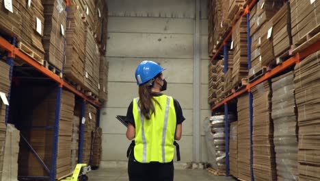 A-female-worker-walking-inside-a-storage-area-of-a-warehouse,-inspecting-the-shelves-while-carrying-a-notepad