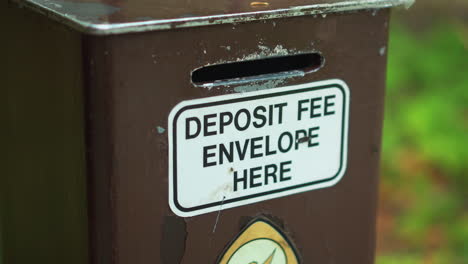 Closeup-of-heavy-duty-outdoor-campground-cash-money-fee-collection-post-with-deposit-envelope-slot-sign-at-camping-site-in-nature