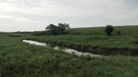 A-quiet,-calm-stream-of-water-in-the-middle-of-a-Kansas-prairie-field-in-the-flints-hills-surrounded-by-green-grass,-a-couple-of-trees-and-hills-in-the-summer-time
