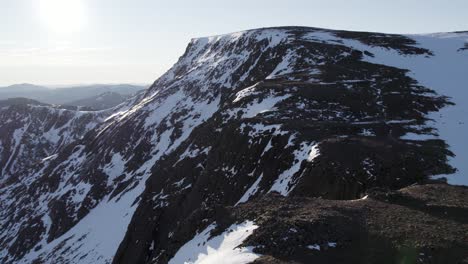 Aerial-drone-footage-rising-above-steep-mountain-cliffs-and-snow-filled-gullies-near-Ben-Macdui-in-the-Cairngorms-National-Park,-Scotland-as-sunshine-reflects-off-of-a-snow-covered-winter-ridge