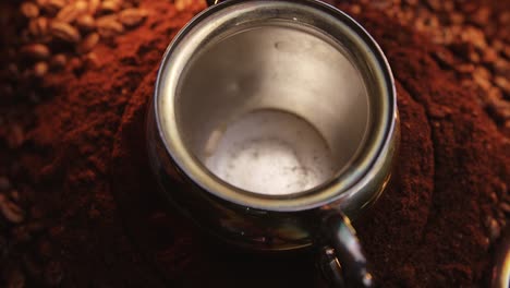 Wirbelnder-Kaffee,-Der-In-Eine-Verzierte-Silberne-Tasse-Strömt,-Die-Sich-Auf-Dunkel-Gemahlenen-Aromatischen-Kaffeebohnen-Dreht