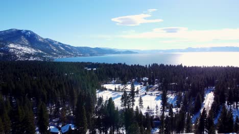 Aerial-drone-shot,-flying-over-the-woods-towards-the-crystal-clear-water-with-the-sun's-reflection-in-Lake-Tahoe,-Nevada-California