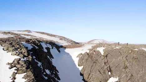 Aerial-drone-footage-reversing-over-a-cliff-face-to-reveal-a-steep-and-dramatic-snow-filled-gully-with-clear-blue-skies-in-the-mountains-near-Ben-Macdui-in-the-Cairngorms-National-Park,-Scotland