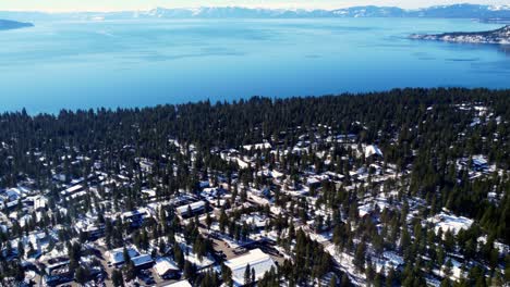 A-beautiful-aerial-drone-shot,-flying-over-buildings-in-the-woods-towards-the-crystal-clear-lake-at-Lake-Tahoe,-Nevada-California