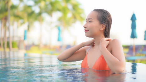 A-Close-up-of-an-attractive-woman-emerging-from-the-swimming-pool-pushes-her-wet-hair-from-her-face,-then-leans-back-along-the-side-of-the-pool