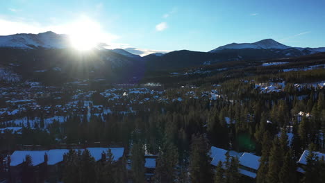 Aerial-shot-of-a-snowy-forest-in-Colorado,-USA-with-the-sun's-rays-being-filtered-by-a-mountain-in-the-background---drone-shot