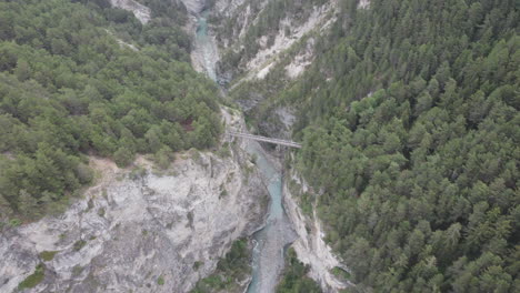 Aerial-drone-shot-flying-over-tilting-down-tracking-a-suspension-bridge-which-joins-two-cliffs-of-a-deep-valley-gorge-together-with-the-Arc-river-flowing-below-in-Aussois,-France