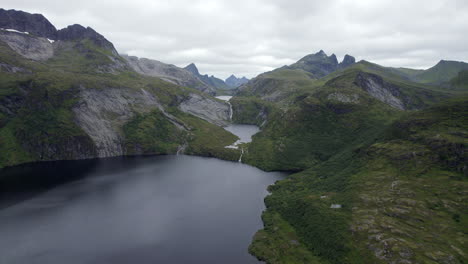 Wide-aerial-shot-of-a-lake-being-fed-by-waterfalls-among-rocky-peaks-and-greenery-in-the-mountains-on-an-overcast-day