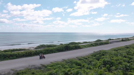 Orbital-drone-shot-of-a-young-couple-driving-a-red-scooter-down-a-highway-along-the-pacific-ocean-coast-on-the-tropical-island-of-Cozumel,-Mexico-shot-in-4k