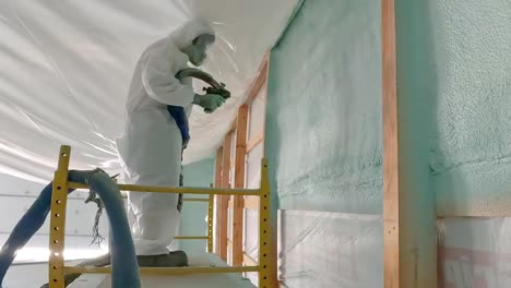 Man-in-disposable-suit-and-respirator-spraying-foam-insulation-onto-an-exterior-wall-of-a-new-construction-while-standing-on-a-scaffold