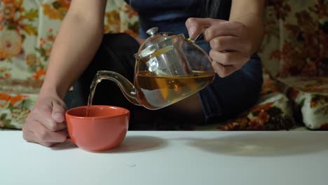 Slow-motion-video:-serving-tea-from-a-transparent-glass-teapot-to-a-brown-colored-cup-indoors