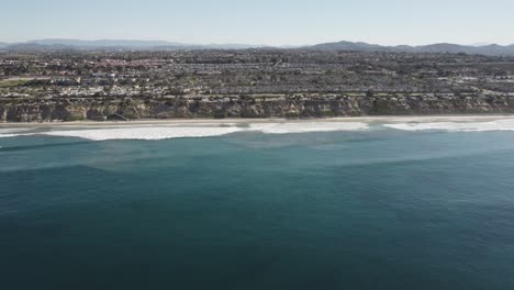 A-beautiful-aerial-drone-shot,-drone-flying-towards-the-coast-over-the-beach-and-a-town-with-mountains-in-the-background,-Carlsbad-State-Beach---California