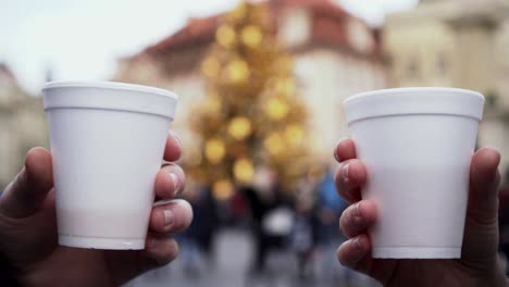 Hands-and-plastic-cups-with-hot-wine-cheering,-christmas-tree,-people-and-blurry-lights-in-background,-closeup-view