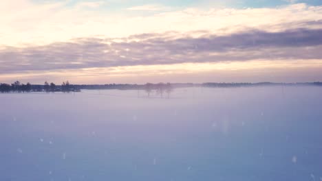 Foggy-winter-rural-landscape-during-snowfall-with-rising-sun-in-horizon,-aerial-view