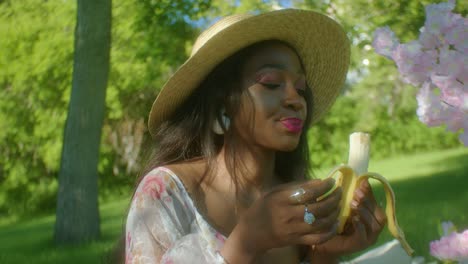 African-woman-wears-hat-and-dress-in-park-on-picnic,-eats-banana-look-around