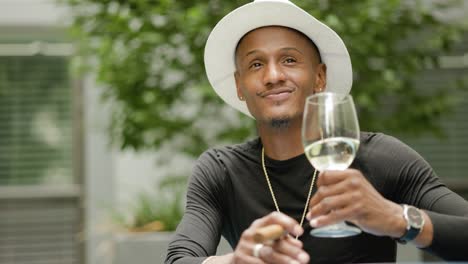 Happy-wealthy-man-wearing-fedora-hat-and-gold-jewelry-sits-at-a-table-swirling-and-drinking-wine-outside-near-green-plants