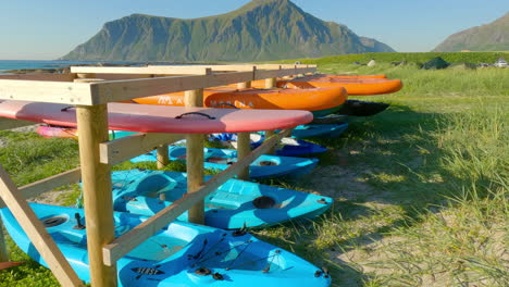 Reverse-handheld-dolly-shot-of-kayaks-and-paddle-boards-on-a-wooden-rack-near-on-Flakstad-beach-in-Lofoten-Norway-with-the-ocean,-mountains-and-green-fields-in-the-background
