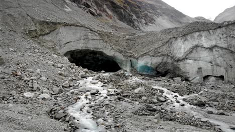 Aerial-flyover-into-the-ice-cave-of-the-Zinal-glacier-in-Valais,-Switzerland-with-a-pan-up-view-from-the-melting-glacial-water-stream-and-crevasses