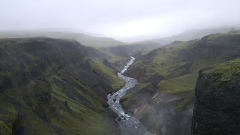 Aerial-push-in-shot-of-a-river-in-the-mountain-gorge-of-Fossárdalur,-in-Iceland,-on-a-cloudy-and-foggy-day