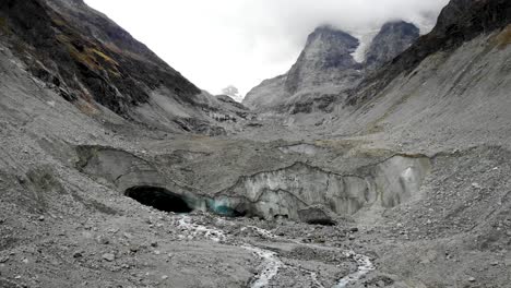 Aerial-flyover-into-the-ice-cave-of-the-Zinal-glacier-in-Valais,-Switzerland-with-a-view-of-the-melting-glacial-water-stream-in-alpine-valley