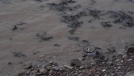 Bicycle-abandoned-in-the-beach,-waves-crushing-into-a-bike-that-was-trowed-into-the-sea,-see-weeds-floating-and-crushing-in-the-beach