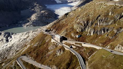Aerial-flyover-over-Furka-mountain-pass-at-the-border-of-Valais-and-Uri-in-Switzerland-with-a-pan-down-view-from-the-Rhone-glacier-down-to-cars-driving-down-the-numerous-turns-of-the-road
