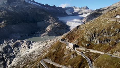 Aerial-flyover-over-Furka-mountain-pass-at-the-border-of-Valais-and-Uri-in-Switzerland-with-a-view-of-the-Rhone-glacier-behind-the-curvy-road-turns-on-a-sunny-summer-day