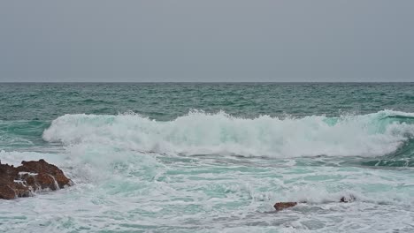 Coast-in-a-bad-weather-day,-gray-sky,-blue-big-waves-crashing-into-orange-rocks-of-the-coastline