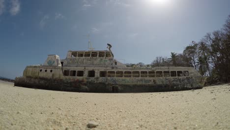 Low-Angle-View-Looking-Up-At-Abandoned-Double-Hulled-Ship-On-Beach-With-Person-Exploring-And-Taking-Photos-On-Roof
