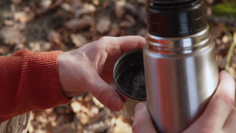 Pouring-hot-tea-or-coffee-from-metal-vacuum-thermos-into-cup,-hands-close-up,-forest-autumn-leaves-in-background